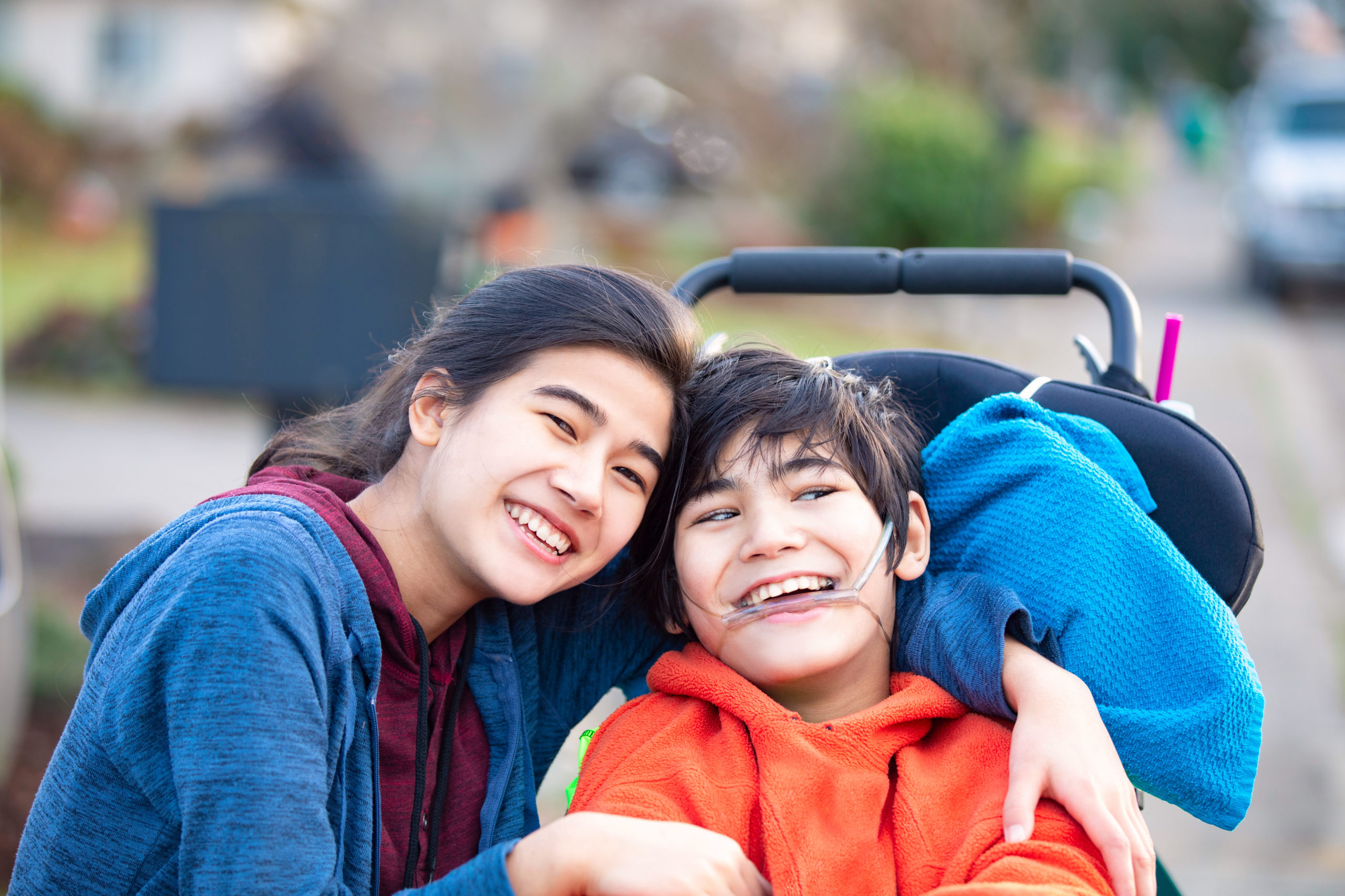 Biracial big sister lovingly hugging disabled little brother in wheelchairoutdoors, smiling