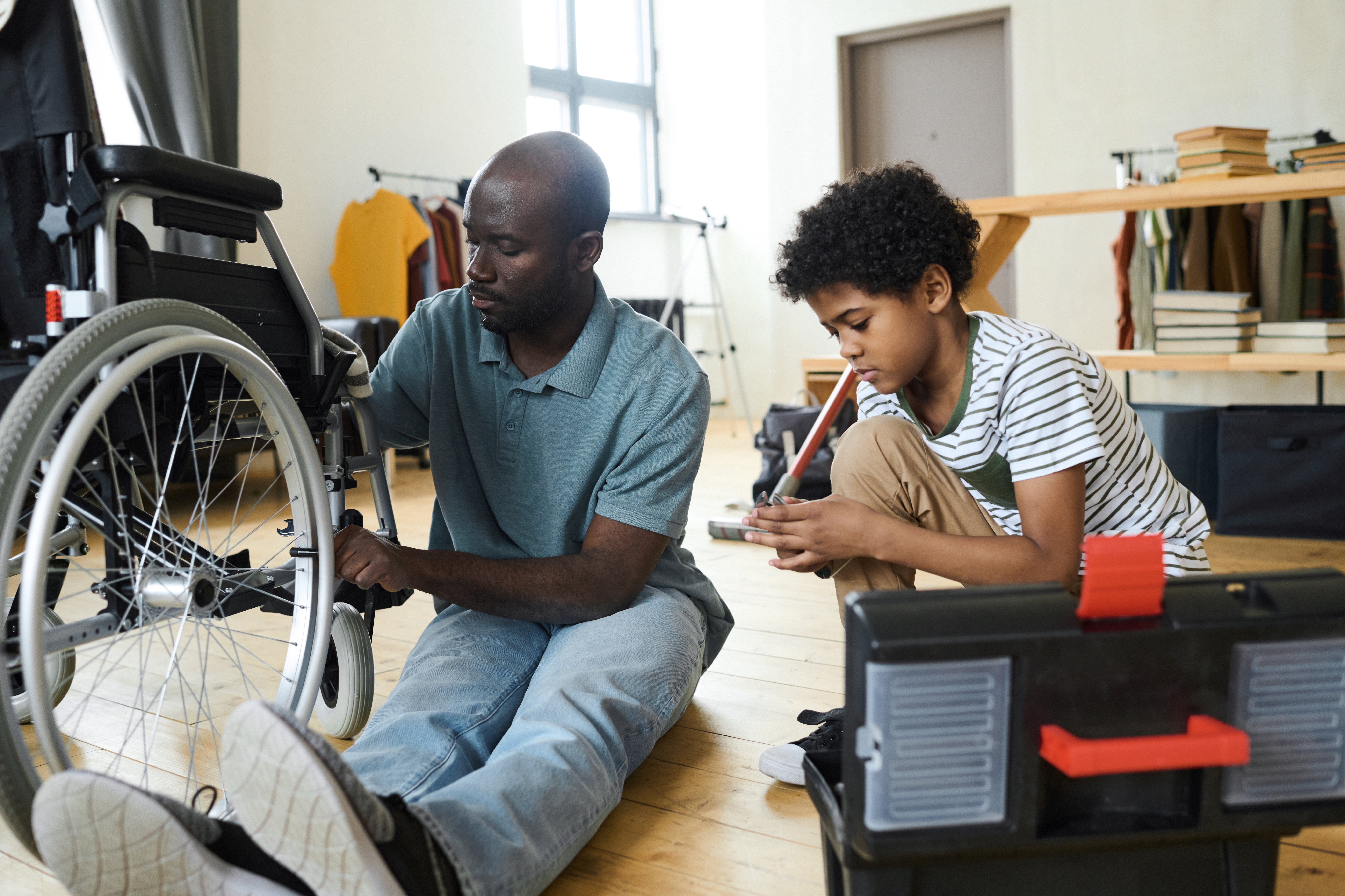 African father with disability sitting on floor and repairing his wheelchair with son helping him and giving tools from box