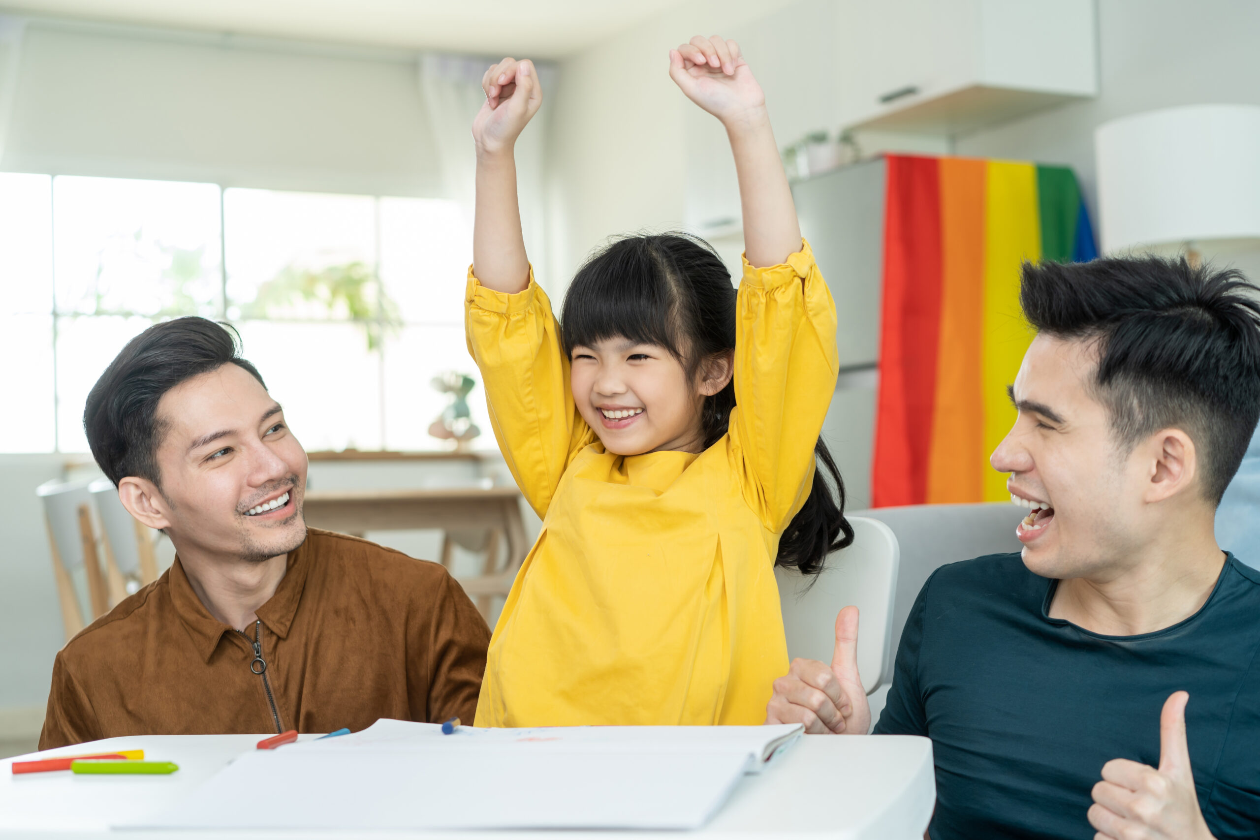 Asian attractive LGBTQ gay family teach young girl kid draw picture. Handsome male couple look at little adorable child daughter play and coloring book in living room, enjoy parenting activity at home