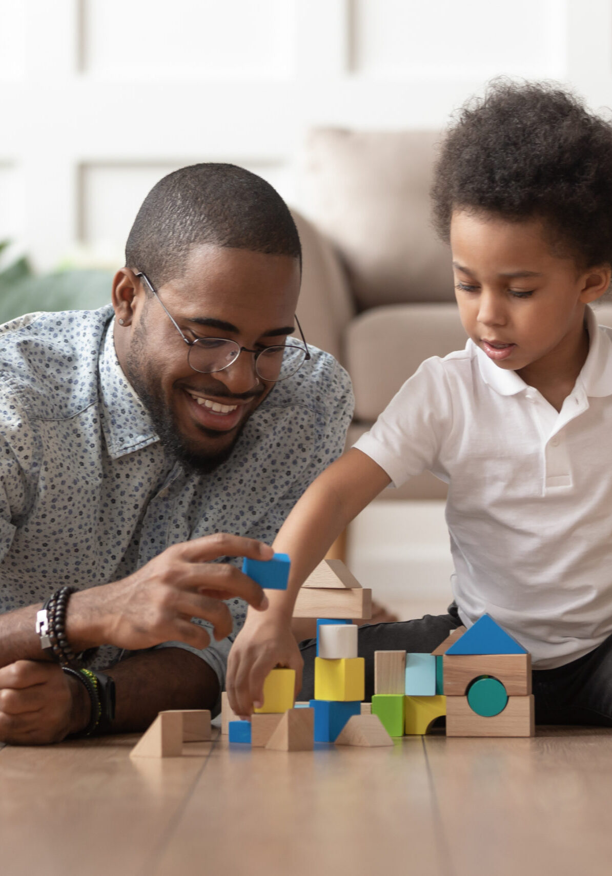 Caring young single black father help cute kid son play on warm floor together, happy african family dad and little child boy having fun building constructor tower from colorful wooden blocks
