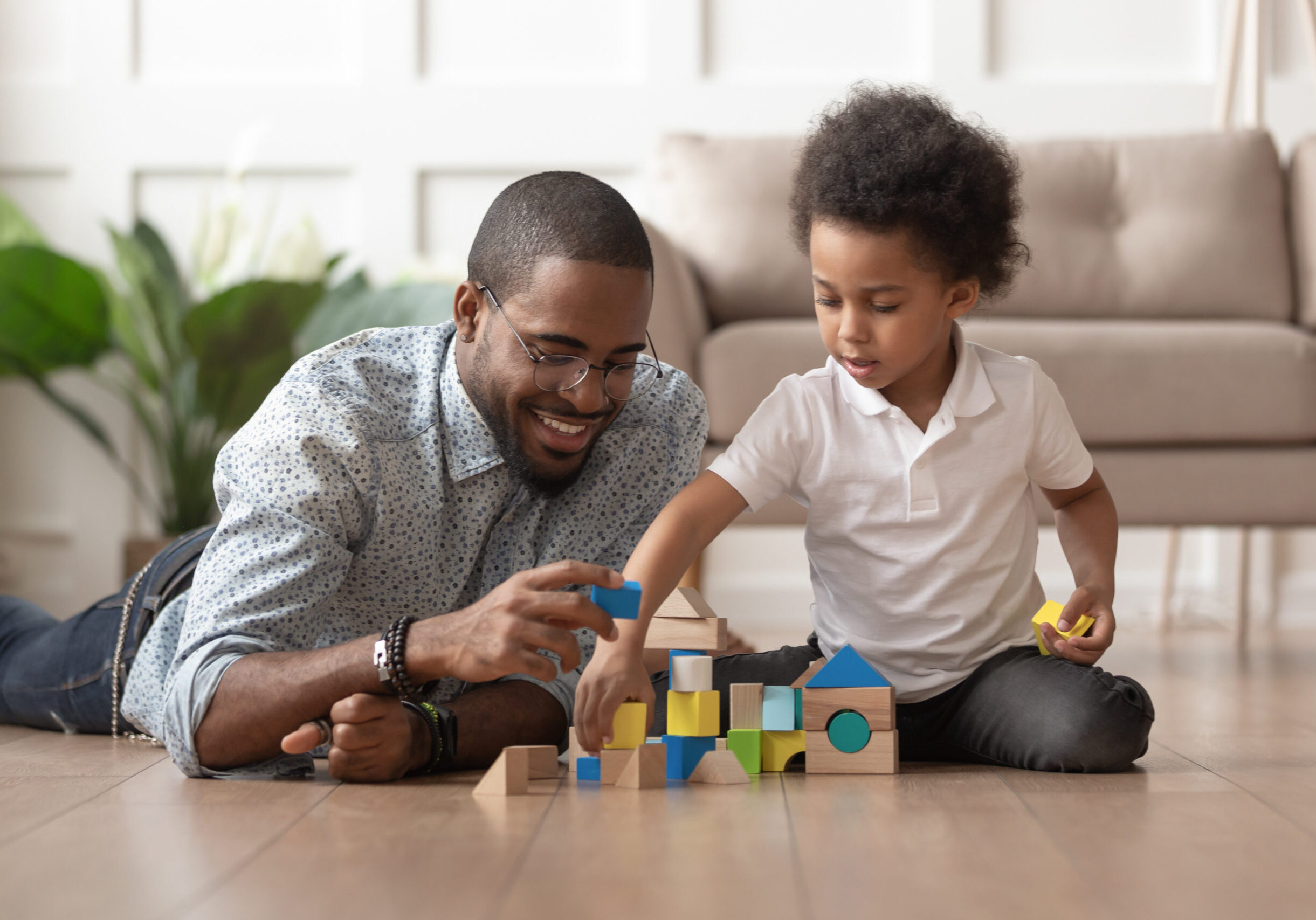Caring young single black father help cute kid son play on warm floor together, happy african family dad and little child boy having fun building constructor tower from colorful wooden blocks