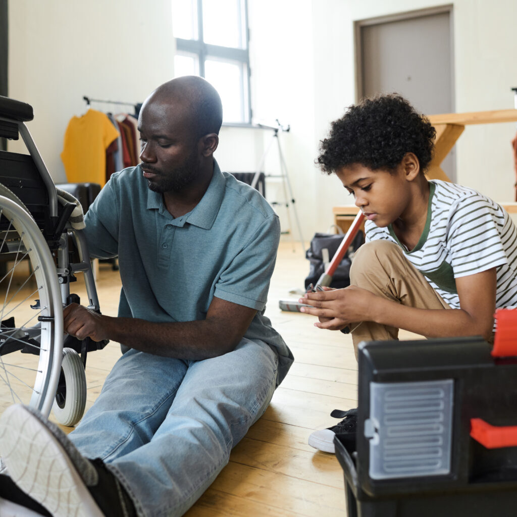 Disabled dad fixing wheelchair with his son African father with disability sitting on floor and repairing his wheelchair with son helping him and giving tools from box